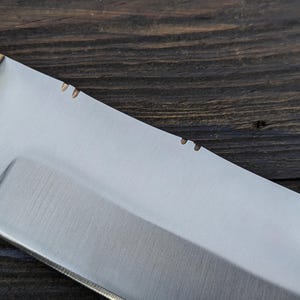 May include: Close-up of a silver knife blade with a wooden handle. The blade has a slight curve and a sharp point. The handle is made of wood and has a light brown color.