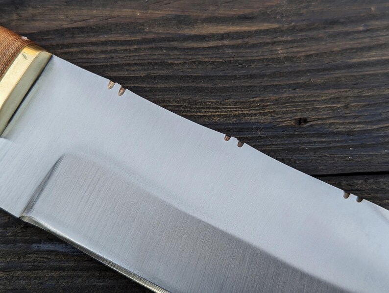 May include: Close-up of a silver knife blade with a wooden handle. The blade has a slight curve and a sharp point. The handle is made of wood and has a light brown color.