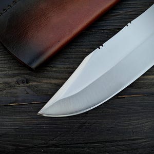 May include: A close-up of a silver knife blade with a serrated edge. The blade is sharp and has a polished finish. The knife is lying on a dark wooden surface.