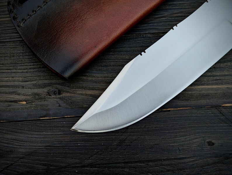 May include: A close-up of a silver knife blade with a serrated edge. The blade is sharp and has a polished finish. The knife is lying on a dark wooden surface.