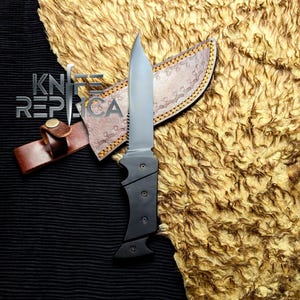 May include: A black and silver knife with a serrated edge and a black handle, resting on a brown leather sheath. The sheath is decorated with a pattern and a leather strap. The knife and sheath are on a textured, golden-brown surface. The text "KNIFE REPLICA" is visible.