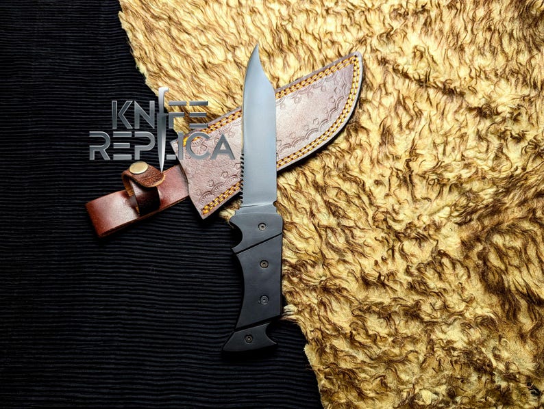 May include: A black and silver knife with a serrated edge and a black handle, resting on a brown leather sheath. The sheath is decorated with a pattern and a leather strap. The knife and sheath are on a textured, golden-brown surface. The text "KNIFE REPLICA" is visible.