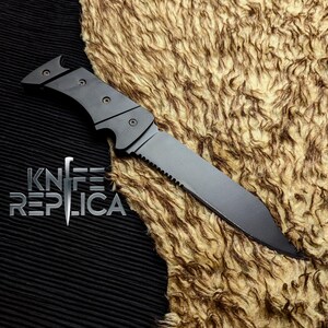 May include: A black folding knife with a serrated edge and a pointed tip. The knife has a matte finish and a handle with three rivets. The text "KNIFE REPLICA" is displayed in silver below the knife.