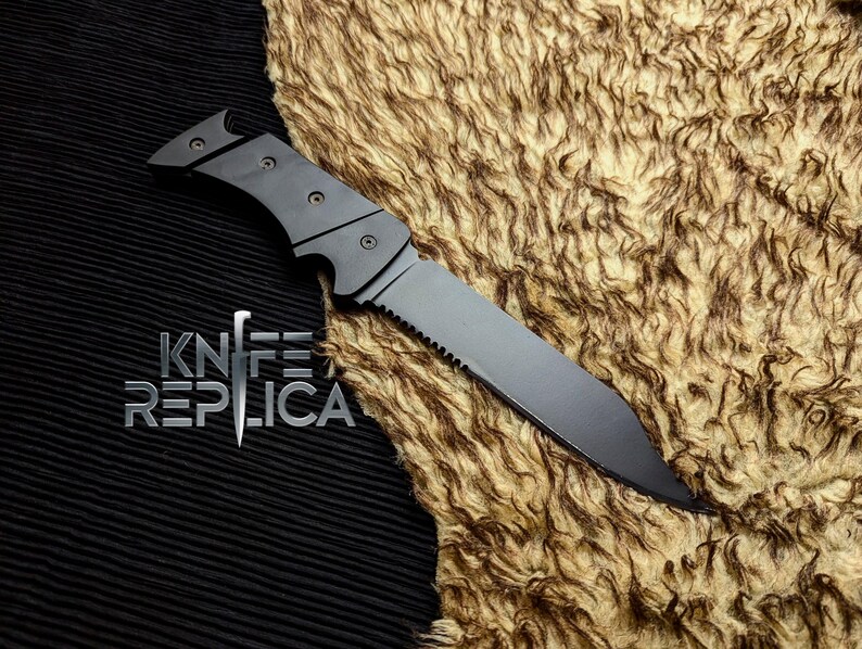 May include: A black folding knife with a serrated edge and a pointed tip. The knife has a matte finish and a handle with three rivets. The text "KNIFE REPLICA" is displayed in silver below the knife.