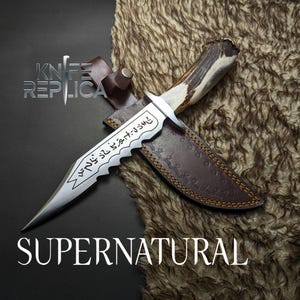 May include: A silver-colored knife with a serrated edge and etched text, resting on a brown leather sheath. The handle is made of a light-colored material with dark brown accents. The word "SUPERNATURAL" is displayed below the knife.