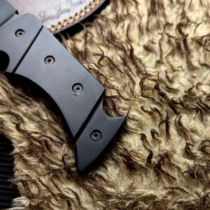 May include: Close-up of a black metal knife with a curved blade and a handle secured by three screws. The knife rests on a textured, brown and beige fur-like material, with a leather sheath visible in the background.