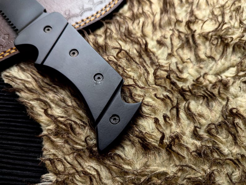 May include: Close-up of a black metal knife with a curved blade and a handle secured by three screws. The knife rests on a textured, brown and beige fur-like material, with a leather sheath visible in the background.
