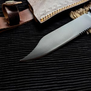 May include: Close-up of a partially serrated, silver-colored knife blade with a black handle. A brown leather sheath and strap are visible, along with a portion of a decorative leather case. The knife is set against a black, textured background.