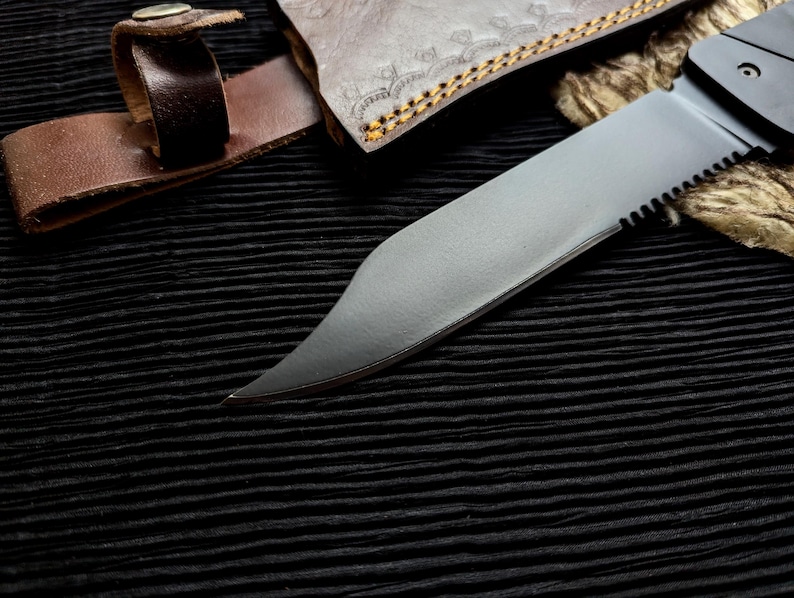 May include: Close-up of a partially serrated, silver-colored knife blade with a black handle. A brown leather sheath and strap are visible, along with a portion of a decorative leather case. The knife is set against a black, textured background.