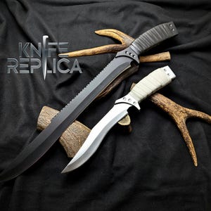 May include: Two knives with black and white handles, with a serrated blade and a straight blade, are displayed on a black fabric background with antlers.