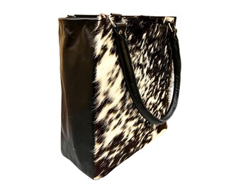 May include: A tote bag with a striking cowhide pattern in black and white. The bag features black leather sides and handles, creating a contrast with the natural hide. The bag is designed for carrying items.