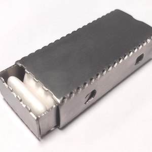 Puede incluir: Una caja rectangular de metal con tapa corredera. La caja está abierta, revelando dos objetos cilíndricos blancos en su interior. Los bordes de la caja y la tapa tienen un diseño decorativo festoneado. La caja es de metal color plata.