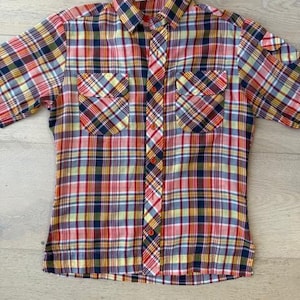 May include: A short-sleeved shirt with a colourful plaid pattern. The shirt features a button-down front and two chest pockets. The plaid design incorporates red, yellow, blue, and green hues, creating a vibrant look.