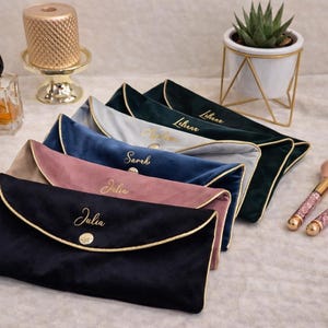 Könnte beinhalten: Eine Auswahl an personalisierten Samt-Clutch-Taschen in verschiedenen Farben, darunter Marineblau, Rosa und Türkis, jeweils mit einem goldgestickten Namen. Make-up-Pinsel und eine kleine Sukkulente sind ebenfalls zu sehen.