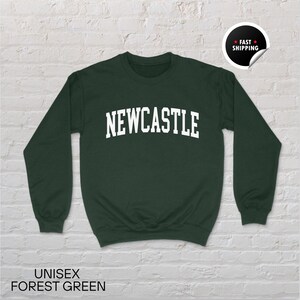 Puede incluir: Sudadera unisex verde bosque con la palabra "NEWCASTLE" en letras blancas. La sudadera tiene mangas largas y cuello redondo. El texto "UNISEX FOREST GREEN" est&aacute; impreso en la parte inferior de la imagen.