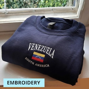 May include: Navy blue sweatshirt with the word "VENEZUELA" embroidered in white above a colorful Venezuelan flag. The words "SOUTH AMERICA" are embroidered below the flag. The word "EMBROIDERY" is in a light blue rectangle at the bottom.
