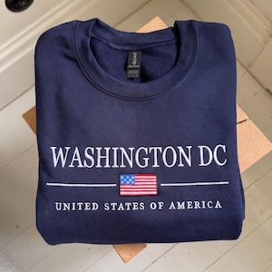 May include: Navy blue sweatshirt with white embroidered text reading "WASHINGTON DC" and "UNITED STATES OF AMERICA". A small American flag patch is centered below the text. The word "EMBROIDERY" is in a light blue box at the bottom.