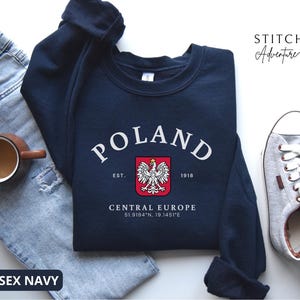 Puede incluir: Una sudadera azul marino de cuello redondo con el texto blanco POLAND en fuente arqueada sobre un escudo rojo con un emblema de águila blanca. Debajo del emblema, el texto dice EST. 1918, CENTRAL EUROPE y las coordenadas 51.9194°N, 19.1451°E.