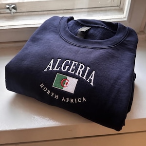 May include: A navy blue crewneck sweatshirt featuring white embroidered text that reads ALGERIA in an arched font above a small embroidered flag of Algeria, with the text NORTH AFRICA embroidered in a smaller white font below the flag.