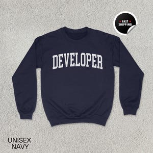 Peut inclure: Sweat-shirt unisexe bleu marine avec le mot "DEVELOPER" en blanc, en lettres arquées. Le sweat-shirt a un col rond et des manches longues. Les mots "UNISEX NAVY" sont imprimés en bas à gauche. Un autocollant "FAST SHIPPING" est en haut à droite.