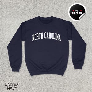 May include: Navy blue unisex sweatshirt with the text "NORTH CAROLINA" in white, arched across the chest. The sweatshirt has a crew neck and long sleeves. A sticker in the upper right corner says "FAST SHIPPING".