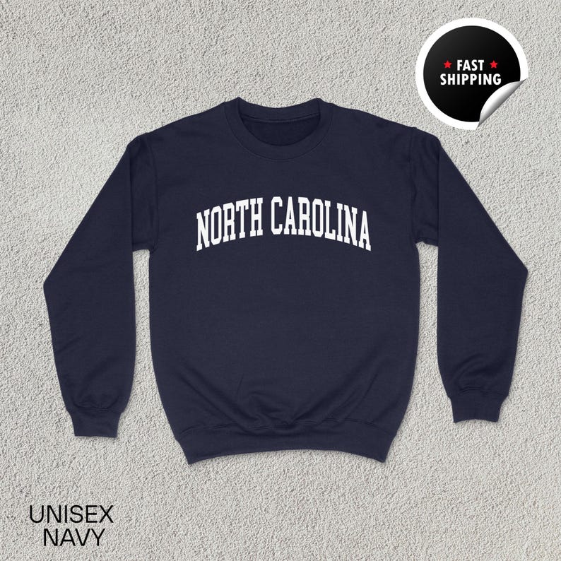 May include: Navy blue unisex sweatshirt with the text "NORTH CAROLINA" in white, arched across the chest. The sweatshirt has a crew neck and long sleeves. A sticker in the upper right corner says "FAST SHIPPING".