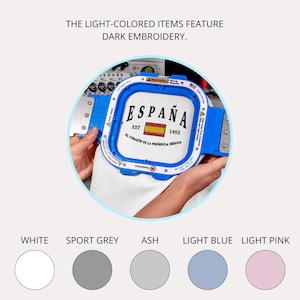 May include: A white fabric is held in a blue embroidery hoop. The fabric features the word "ESPA&Ntilde;A" with a Spanish flag and the text "EST 1492". The text "EL CORAZ&Oacute;N DE LA PEN&Iacute;NSULA IB&Eacute;RICA" is also visible. Color swatches of white, sport grey, ash, light blue, and light pink are shown.