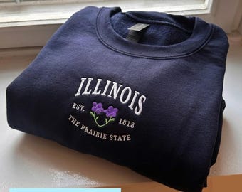 May include: A navy blue sweatshirt with white embroidered text reading "ILLINOIS" above a floral design and the text "EST. 1818 THE PRAIRIE STATE". The word "EMBROIDERY" is in a light blue box at the bottom.
