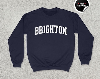 Könnte beinhalten: Marineblaues Sweatshirt mit dem Wort "BRIGHTON" in weißer, bogenförmiger Schrift. Das Sweatshirt hat einen Rundhalsausschnitt und lange Ärmel. Ein Aufkleber in der oberen rechten Ecke zeigt "FAST SHIPPING". Die Worte "UNISEX NAVY" sind unten aufgedruckt.