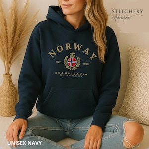 以下が含まれることがあります： ネイビーブルーのフード付きスウェットシャツ。アーチ状のゴールドのフォントでNORWAYと書かれ、その下にノルウェー国旗、EST 1905、SCANDINAVIA、座標59.9139 N, 10.7522 Eがゴールドで描かれた円形のエンブレムが配置されています。