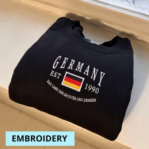 May include: Black sweatshirt with white embroidered text and a German flag design. The text reads "GERMANY EST 1990" with the phrase "DAS LAND DER DICHTER UND DENKER" below. The word "EMBROIDERY" is in a light blue rectangle.