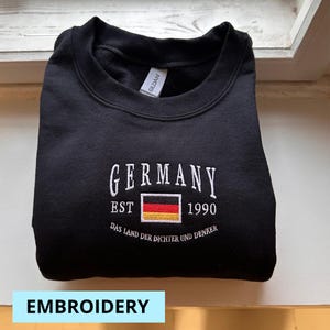 May include: Black crewneck sweatshirt with white embroidered text "GERMANY" above a German flag graphic and the year "1990". The phrase "DAS LAND DER DICHTER UND DENKER" is embroidered below. The word "EMBROIDERY" is in a light blue rectangle at the bottom.