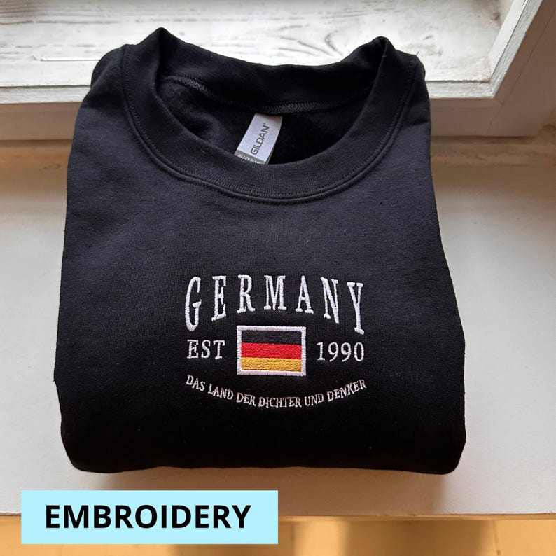 May include: Black crewneck sweatshirt with white embroidered text "GERMANY" above a German flag graphic and the year "1990". The phrase "DAS LAND DER DICHTER UND DENKER" is embroidered below. The word "EMBROIDERY" is in a light blue rectangle at the bottom.