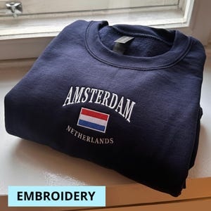 Może przedstawiać: Granatowa bluza z haftowanym białym napisem "AMSTERDAM", nad małą flagą Holandii i słowem "NETHERLANDS". Bluza jest złożona i leży na białej powierzchni.