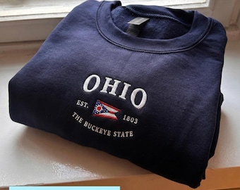 May include: Navy blue sweatshirt with white embroidered text reading "OHIO" above a small Ohio state flag and the text "EST. 1803 THE BUCKEYE STATE". The word "EMBROIDERY" is in a light blue rectangle at the bottom.