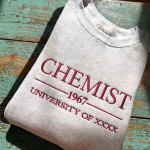 May include: A light gray sweatshirt with the word "CHEMIST" embroidered in burgundy thread. Below the word "CHEMIST" is the year "1967" and "UNIVERSITY OF XXXX". The top left corner has a label that says "CUSTOM EMBROIDERED SHIRT".