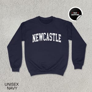 Puede incluir: Sudadera unisex azul marino con la palabra "NEWCASTLE" en letras blancas en el pecho. La sudadera tiene cuello redondo y mangas largas. Una pegatina de "Env&iacute;o r&aacute;pido" est&aacute; en la esquina superior derecha. Las palabras "UNISEX NAVY" est&aacute;n en la parte inferior.