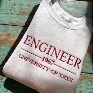 May include: A light gray sweatshirt with the word "ENGINEER" embroidered in burgundy, along with "1967" and "UNIVERSITY OF XXXX". The top left corner has a maroon tag that says "CUSTOM EMBROIDERED SHIRT".