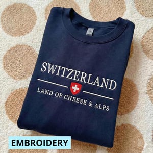 以下が含まれることがあります： ネイビーブルーのスウェットシャツ。白で「SWITZERLAND」と刺繍され、スイス国旗のエンブレムと「LAND OF CHEESE & ALPS」の文字が施されています。「EMBROIDERY」の文字が下にあります。