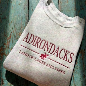 May include: A light gray sweatshirt with the word "ADIRONDACKS" embroidered in burgundy, along with "LAND OF LAKES AND PINES" and a moose graphic. The sweatshirt is folded on a weathered, teal-colored wooden surface.