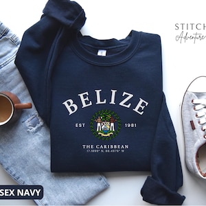 May include: A navy blue crewneck sweatshirt featuring the white text BELIZE in an arched font, the national coat of arms, the text EST 1981, THE CARIBBEAN, and the coordinates 17.1899 N, 88.4976 W. The sweatshirt is displayed with light blue jeans and trainers.