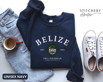 May include: A navy blue crewneck sweatshirt featuring the white text BELIZE in an arched font, the national coat of arms, the text EST 1981, THE CARIBBEAN, and the coordinates 17.1899 N, 88.4976 W. The sweatshirt is displayed with light blue jeans and sneakers.