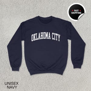 Puede incluir: Sudadera azul marino con el texto blanco "OKLAHOMA CITY" en letras arqueadas. La sudadera tiene cuello redondo y mangas largas. Las palabras "UNISEX NAVY" están impresas en la parte inferior izquierda. Una pegatina de "FAST SHIPPING" está en la esquina superior derecha.