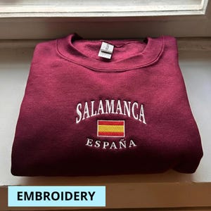 Może przedstawiać: Bordowa bluza z haftowanym napisem "SALAMANCA ESPAÑA" i hiszpańską flagą. Bluza z okrągłym dekoltem jest złożona. Słowo "EMBROIDERY" znajduje się na dole obrazu.