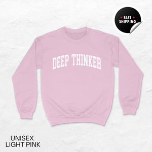 May include: A light pink unisex sweatshirt with the text "DEEP THINKER" in white block letters across the chest. The sweatshirt has long sleeves and a crew neck. The words "UNISEX LIGHT PINK" are at the bottom.