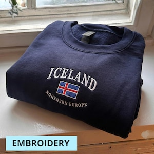 Puede incluir: Sudadera azul marino con la palabra "ICELAND" bordada en blanco, junto con la bandera islandesa. El texto "NORTHERN EUROPE" est&aacute; bordado debajo. La palabra "EMBROIDERY" est&aacute; en un cuadro azul claro en la parte inferior.