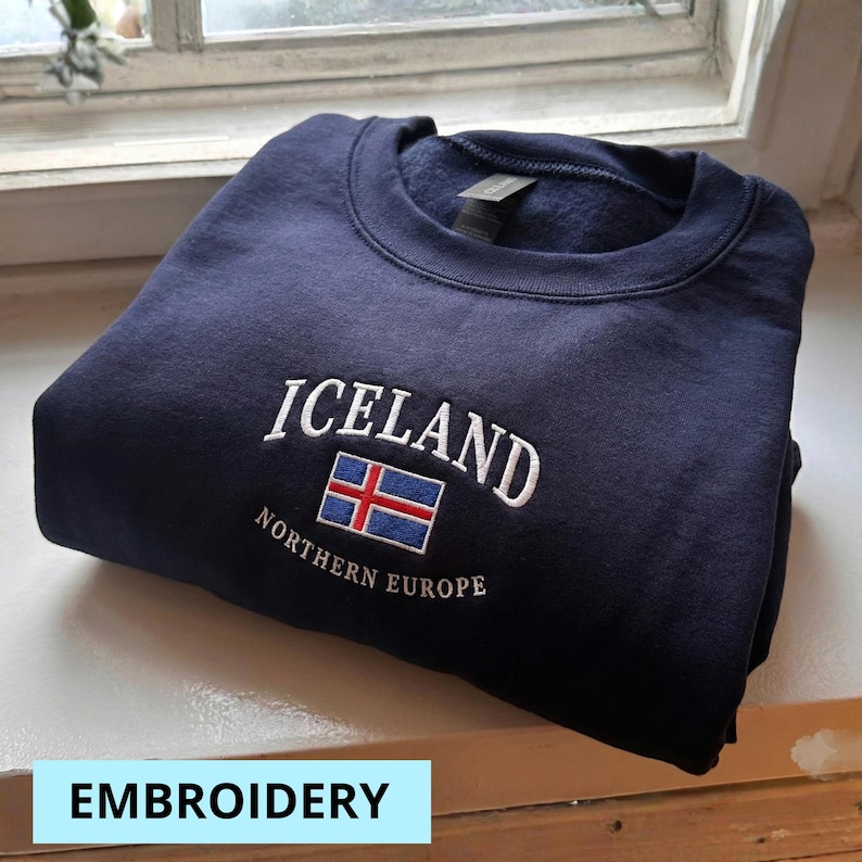 Puede incluir: Sudadera azul marino con la palabra "ICELAND" bordada en blanco, junto con la bandera islandesa. El texto "NORTHERN EUROPE" est&aacute; bordado debajo. La palabra "EMBROIDERY" est&aacute; en un cuadro azul claro en la parte inferior.