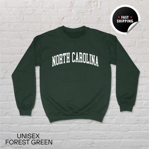 May include: A forest green unisex sweatshirt with the text "NORTH CAROLINA" in white, arched across the chest. The sweatshirt has long sleeves and a crew neck. The words "UNISEX FOREST GREEN" are at the bottom.