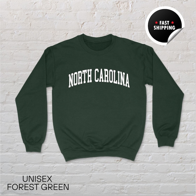 May include: A forest green unisex sweatshirt with the text "NORTH CAROLINA" in white, arched across the chest. The sweatshirt has long sleeves and a crew neck. The words "UNISEX FOREST GREEN" are at the bottom.