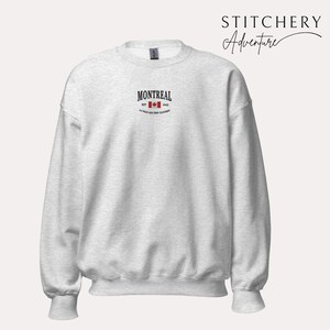 May include: A light gray sweatshirt with the word "MONTREAL" in black text above a red maple leaf and the text "EST. 1642". The sweatshirt has a crew neck and long sleeves. The brand name "STITCHERY Adventure" is in the upper right corner.
